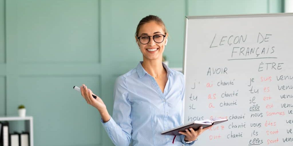 Friendly happy teacher recommending foreign languages school, giving French lesson, standing near whiteboard and smiling at camera. Positive young tutor satisfied with educational services