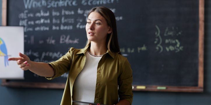 Medium shot of female English teacher holding tablet computer addressing student with question, while discussing new grammar rules in group class against blackboard at language school